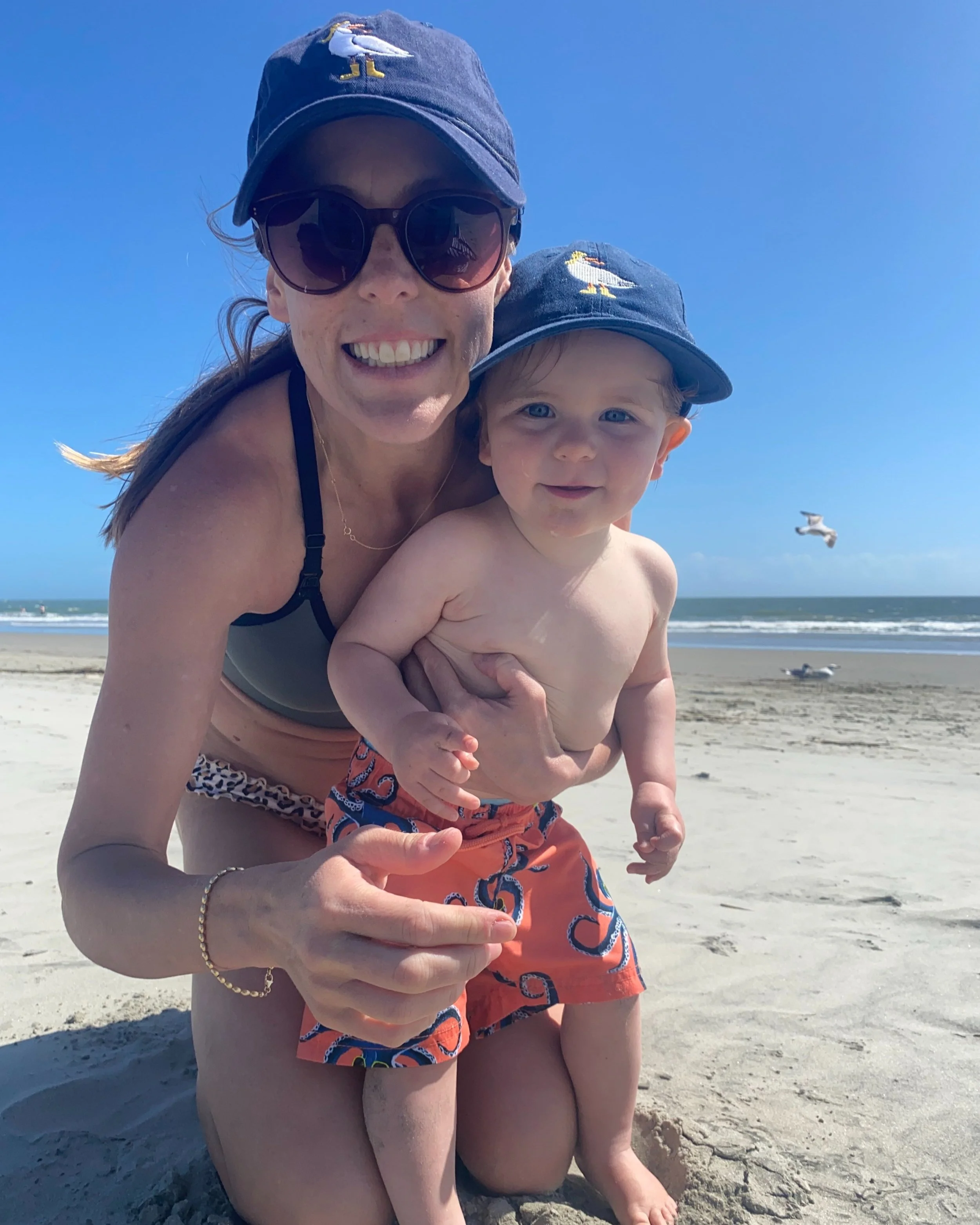 Woman kneels in the sand, at the beach, and holds toddler
